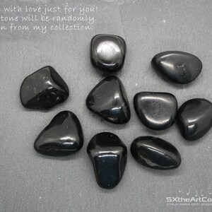 May include: A collection of nine smooth, black stones. The stones are tumbled and polished, and they have a glossy finish. The stones are arranged on a gray surface. The text "Made with love just for you! The stone will be randomly chosen from my collection." is written above the stones. The text "SXtheArtConcept STALO XINAFRI ART DESIGN JEWELRY" is written below the stones.