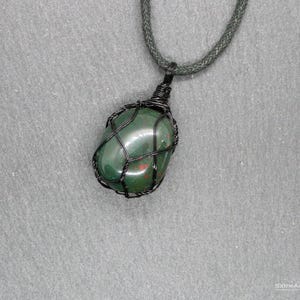 Op de afbeelding: Een groene edelsteen hanger ketting. De gepolijste steen is ingekapseld in een zwarte draadkooi en hangt aan een donkergroen koord. De steen heeft rode vlekken tegen een grijze achtergrond.