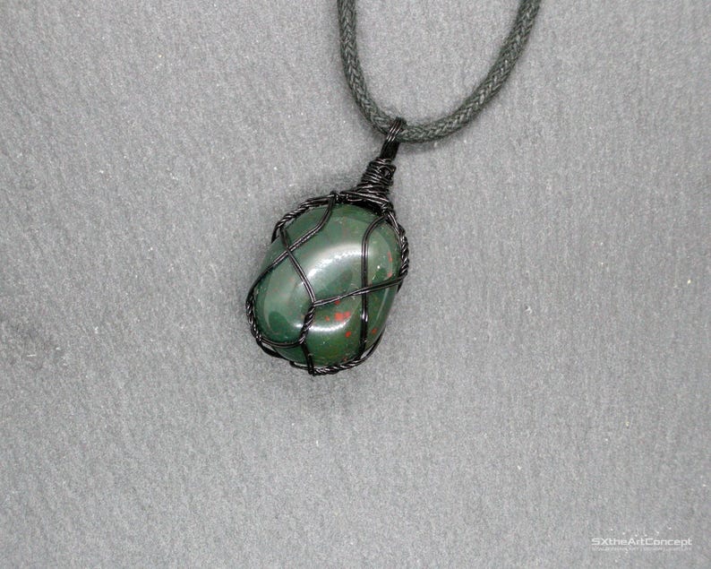 Op de afbeelding: Een groene edelsteen hanger ketting. De gepolijste steen is ingekapseld in een zwarte draadkooi en hangt aan een donkergroen koord. De steen heeft rode vlekken tegen een grijze achtergrond.