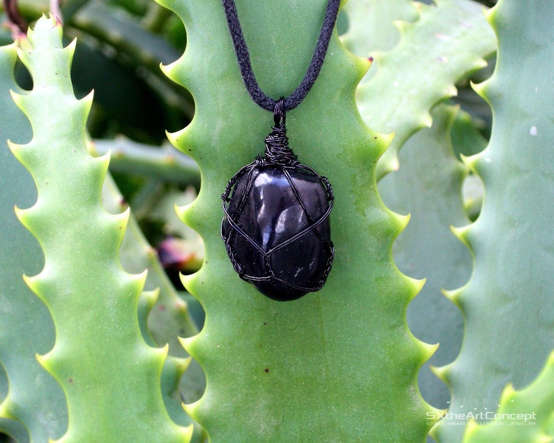 May include: Black onyx gemstone pendant wrapped in black wire, hanging from a black cord necklace. The pendant is displayed against a green leafy background.