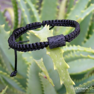 Pode incluir: Pulseira tran&ccedil;ada preta com uma conta de lava preta. A pulseira est&aacute; em uma planta verde.