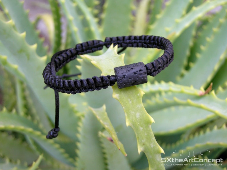 Pode incluir: Pulseira tran&ccedil;ada preta com uma conta de lava preta. A pulseira est&aacute; em uma planta verde.