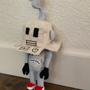 May include: A handmade felt robot doll with a beige head and body, gray arms and legs, and black hands. The robot has a curved antenna, black eyes, and a mouth. It wears red shoes with white soles. The robot's chest has a control panel design.