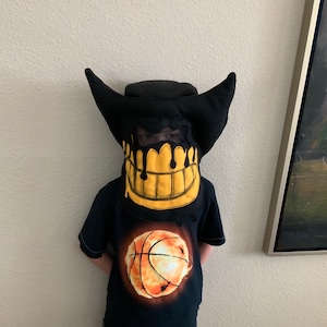 May include: A child wearing a black t-shirt with a basketball graphic and a black felt mask with a yellow dripping smile. The mask is shaped like a cartoon character's head with large ears.