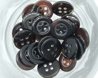 以下が含まれることがあります： ガラスのボウルに、茶色と黒のボタンが詰められています。ボタンはそれぞれ4つの穴があり、ランダムなパターンで配置されています。