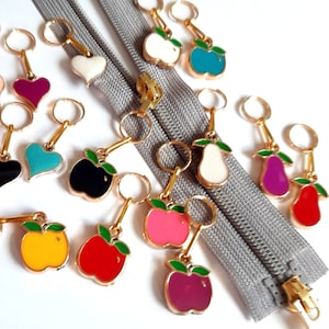 Könnte beinhalten: Ein Set mit 16 bunten Maschenmarkierern mit Herz- und Fruchtmotiven. Die Markierer sind aus Metall mit goldener Oberfläche und haben eine Schlaufe zum Befestigen an Stricknadeln. Die Markierer sind auf einer weißen Oberfläche mit einem grauen Reißverschluss angeordnet.