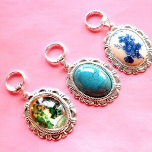 Op de afbeelding: Drie zilveren sleutelhangers met verschillende ovale bedels. De bedels hebben een blauw en wit bloemenmotief, een turquoise steen en een vintage afbeelding van een vrouw.