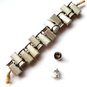 Könnte beinhalten: Ein silbernes Armband aus miteinander verbundenen Patronenhülsen mit einer Schnur an einem Ende. Das Armband hat zwei silberne Verschlüsse, einen mit einer Feder und einen mit einem Loch.