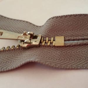 May include: A close-up of a beige zipper with a metal slider and pull tab. The zipper is open and the teeth are visible.