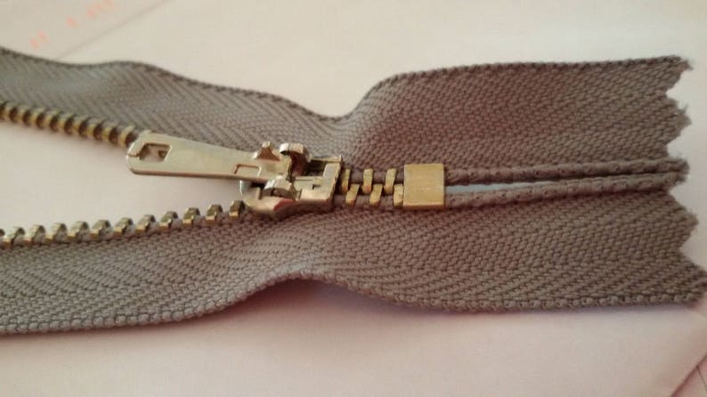 May include: A close-up of a beige zipper with a metal slider and pull tab. The zipper is open and the teeth are visible.