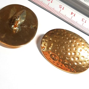 Könnte beinhalten: Zwei goldfarbene, oval geformte Knöpfe. Ein Knopf hat auf der Rückseite eine Schlaufe zum Annähen. Der andere Knopf hat eine strukturierte Oberfläche mit kleinen, gleichmäßig verteilten Vertiefungen. Ein Lineal ist im Hintergrund.