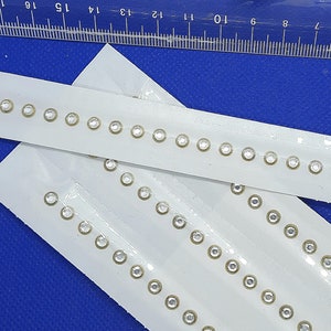 May include: White ribbon trim with rows of clear rhinestones. The trim is on a blue background.