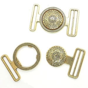 Könnte beinhalten: Zwei goldfarbene Metallgürtelschnallen mit verzierten Blumendesigns. Jede Schnalle hat einen T-förmigen Bügel mit einem runden Dekorationsstück in der Mitte.