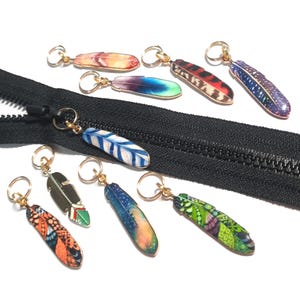 Può includere: Una collezione di tiretti per cerniere a forma di piuma colorata con anelli dorati. Le piume presentano vari motivi e colori, tra cui blu, arancione, verde e viola. È presente anche una cerniera nera.