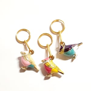 May include: Three gold stitch markers with colorful bird charms. The birds are painted in pink, yellow, and blue with white accents.