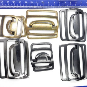 Könnte beinhalten: Sechs Metall-Gürtelschnallen, zwei sind golden, zwei sind schwarz und zwei sind silber. Die Schnallen sind alle gleich groß und gleich geformt und haben ein schlichtes Design.