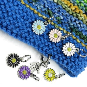 Könnte beinhalten: Ein Set mit sieben bunten Daisy-Maschenmarkierern zum Stricken oder Häkeln. Die Markierer sind aus Metall und haben einen federbelasteten Verschluss. Die Markierer sind in verschiedenen Farben, darunter Schwarz, Lila, Gelb, Rosa und Blau. Die Markierer sind auf einem blauen gestrickten Stoff angeordnet.