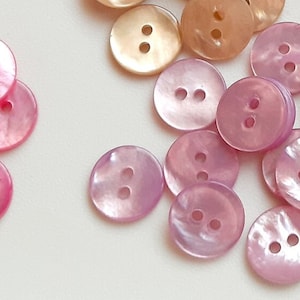 以下が含まれることがあります： 2つの穴のあるピンクと白の虹色のボタンのコレクション。ボタンは白い表面にランダムなパターンで配置されています。