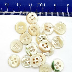 Könnte beinhalten: Eine Sammlung von 15 weißen Perlmuttknöpfen mit jeweils vier Löchern. Die Knöpfe sind rund und haben einen leichten irisierenden Glanz.