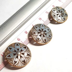 May include: Three round decorative buttons with a floral design. The buttons have a gold-coloured rim and a textured, silver-coloured centre. The floral design features light blue and silver petals. A ruler is in the background.