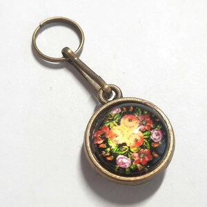 May include: A circular keyring featuring a floral design. The keyring has a bronze-toned metal frame and a clear, domed cover over the floral image. The flowers are in shades of red, orange, yellow, and pink against a black background.