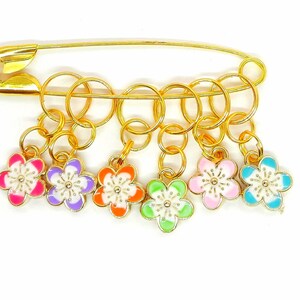 Könnte beinhalten: Ein Satz von sechs goldenen Maschenmarkierern mit bunten Blumenanhängern. Die Marker sind an einer goldenen Sicherheitsnadel befestigt.