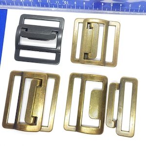 Könnte beinhalten: Vier Metall-Gürtelschnallen, eine schwarze und drei goldene. Die goldenen Schnallen haben eine strukturierte Oberfläche und sind in leicht unterschiedlichen Formen.