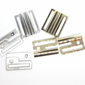 Op de afbeelding: Een assortiment metalen bh-bandjes hardware in zilver- en goudtinten. De hardware omvat rechthoekige schuifregelaars en haak-en-oogsluitingen, ideaal voor lingerie en kleding maken. De stukken zijn gerangschikt op een witte ondergrond.