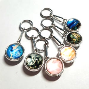 May include: A selection of keyrings with decorative, circular pendants. Each keyring has a silver-coloured metal ring and a matching pendant with a unique, colourful design. The pendants display various abstract patterns and images.