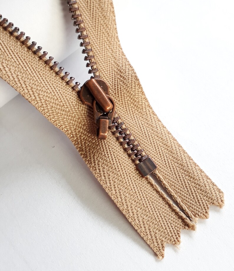 May include: A close-up of a brown zipper with a copper slider. The zipper is open and the teeth are visible.