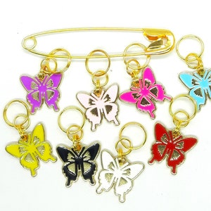 Könnte beinhalten: Eine Sammlung bunter Schmetterlingsanhänger in verschiedenen Farben, darunter Lila, Rosa und Blau, an goldfarbenen Ringen befestigt. Oben befindet sich eine große goldene Sicherheitsnadel. Die Anhänger sind wahrscheinlich für Schmuck- oder Bastelprojekte.