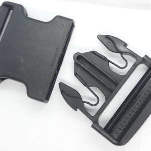 May include: A black plastic side-release buckle, unfastened, against a white background. The buckle consists of two parts: a male and a female component. The female part has a series of teeth for secure fastening. The male part has the text "21 PAT. PEND."