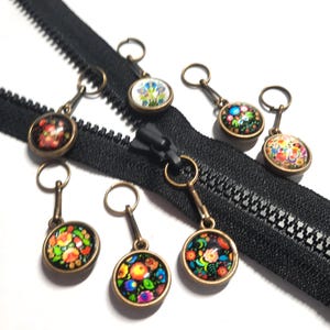 May include: A black zip fastener with multiple decorative zipper pulls. The pulls feature colourful floral designs set in bronze-coloured metal. The zip is open, and the pulls are attached to the slider.