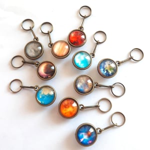May include: A selection of bronze keyrings, each with a circular glass pendant depicting a planet. The planets showcase diverse colors such as red, blue, orange, and grey, with detailed planetary surfaces. The keyrings are arranged on a white background.