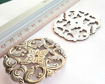 May include: Two silver-coloured metal buckles with detailed floral and scrollwork patterns. A ruler with centimetre measurements is in the background. The buckles have a vintage or antique appearance, suggesting they are for decorative use.