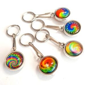 Może przedstawiać: Kolekcja breloków do kluczy z wzorem tie-dye. Każdy brelok ma okrągły, kolorowy wzór tie-dye, zamknięty w srebrnej metalowej oprawie. Breloki są przymocowane do srebrnego metalowego pierścienia.