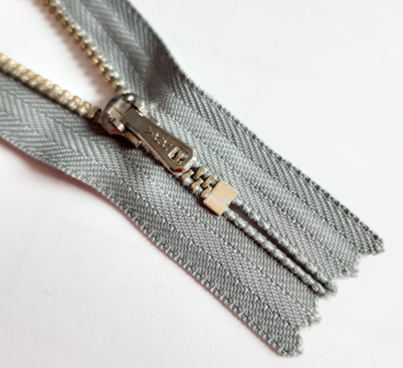May include: A silver zipper with a silver slider and a gray fabric tape. The zipper is open and the slider is in the middle of the tape.