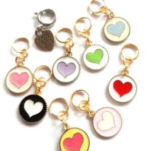 Könnte beinhalten: Acht goldene Maschenmarkierer mit Herzen in verschiedenen Farben. Die Herzen sind rosa, lila, grün, blau, rot, weiß, gelb und schwarz. Jeder Marker hat einen goldenen Ring und einen kleinen Anhänger mit dem Text "Hand Made With Love".