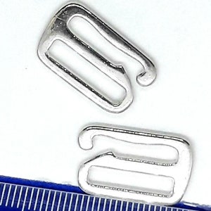 Op de afbeelding: Twee zilveren metalen bh-bandverstellers. De verstellers zijn achtvormig met een klein haakje aan het ene uiteinde.