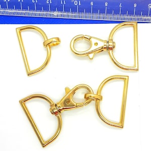 May include: Three gold-toned metal hardware pieces for attaching to bags, straps, or other accessories. The pieces include a swivel hook, a D-ring, and a smaller D-ring.
