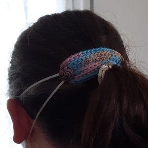Op de afbeelding: Een gebreid hoofdband in roze, blauw en bruine tinten. Het hoofdband heeft een knoop met een swirl-ontwerp erop. Het hoofdband wordt gebruikt om een ​​gezichtsmasker op zijn plaats te houden.