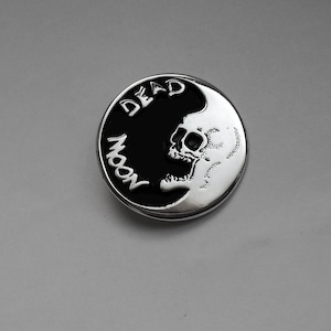 Può includere: Un emblema rotondo in metallo color argento con un design in bianco e nero. Il design presenta un teschio sul lato destro e le parole "DEAD MOON" in lettere bianche sul lato sinistro. L'emblema ha un bordo rialzato.