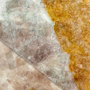 May include: Close-up of several translucent mica sheets in shades of brown, gold, and gray. The sheets have a layered, flaky appearance with visible crystalline structures. The overall effect is delicate and textured.