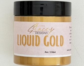 May include: A gold-coloured jar of "Liquid Gold" paint by Stacey Designs, with a black lid and a pink label. The label displays "Liquid Gold" in gold lettering, and the size is 118ml. The jar is a craft supply.