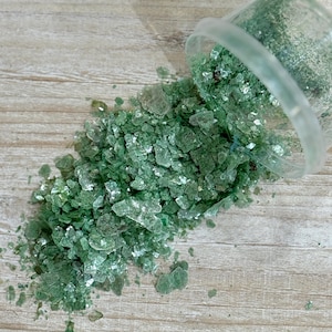 May include: Green mica flakes spilling from a clear plastic container onto a wooden surface. The flakes have a shimmering, translucent quality, with varying shades of green. The container is partially visible, tilted to the side.
