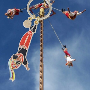 Puede incluir: Un llavero dorado con un colgante que representa a una persona con un traje rojo y negro, colgada boca abajo. El fondo muestra un cielo azul y personas realizando una danza tradicional. El llavero tiene un pequeño dije circular.