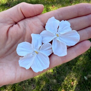 Peut inclure: Deux boucles d'oreilles en forme de fleur blanche avec des pétales texturés et de petites perles dorées au centre de chaque fleur. Le design floral présente cinq pétales distincts par pièce avec une finition mate et une forme organique.
