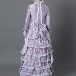 May include: A lavender Victorian-style dress with multiple ruffled layers and white lace trim. The long-sleeved dress features a high collar, a fitted bodice, and a matching hat with a purple ribbon. The dress is made of a shiny fabric.