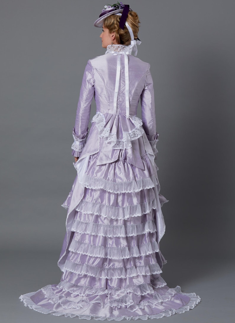 May include: A lavender Victorian-style dress with multiple ruffled layers and white lace trim. The long-sleeved dress features a high collar, a fitted bodice, and a matching hat with a purple ribbon. The dress is made of a shiny fabric.
