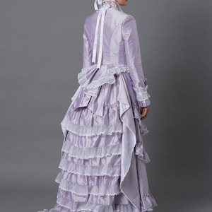 May include: A lavender Victorian-style dress with multiple ruffled layers and white lace trim. The long-sleeved dress features a high collar, a small hat with floral accents, and a long train. The dress is made of a shiny fabric.